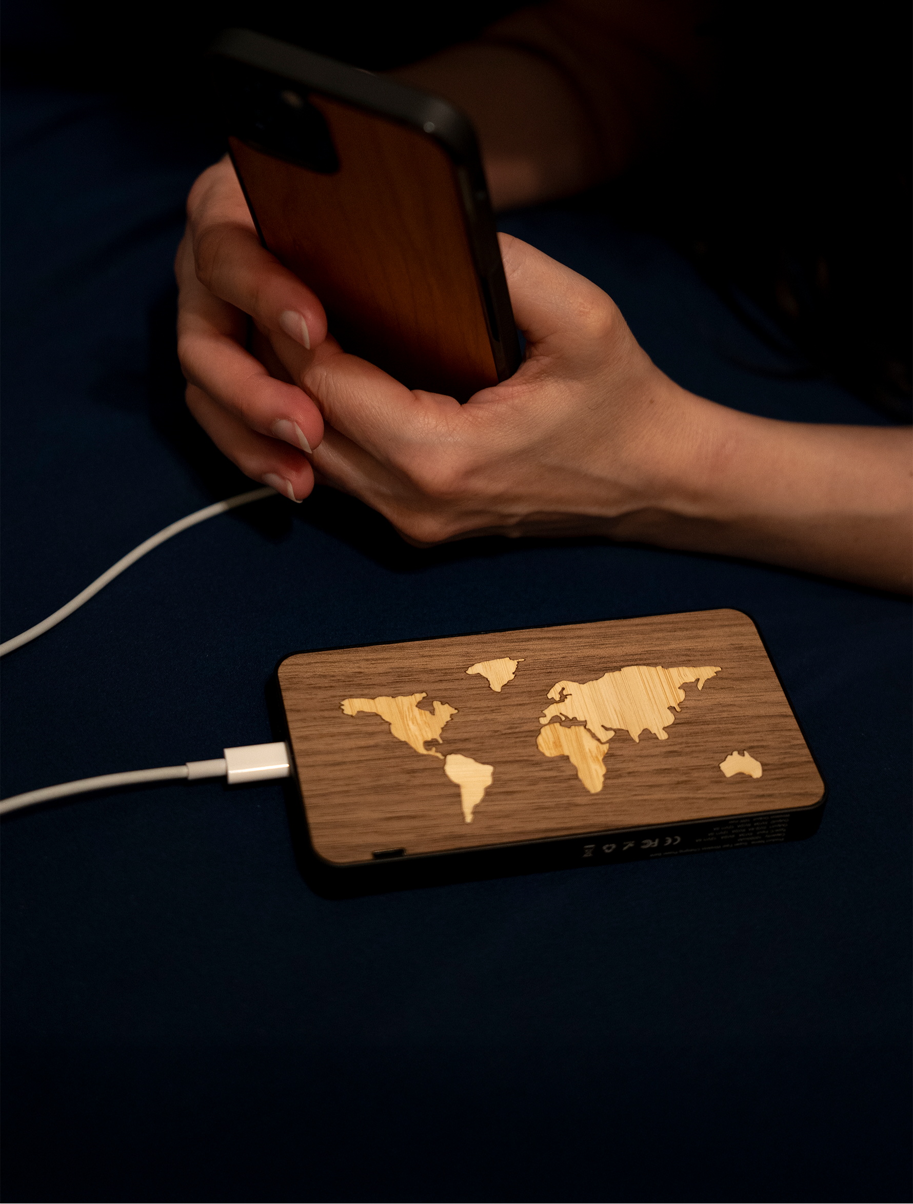 Mains d’une femme tenant un iPhone 12 Pro avec une coque en bois "Le Bois", relié par un câble USB à la batterie portable en bois "La Mappemonde" de 10 000 mAh.