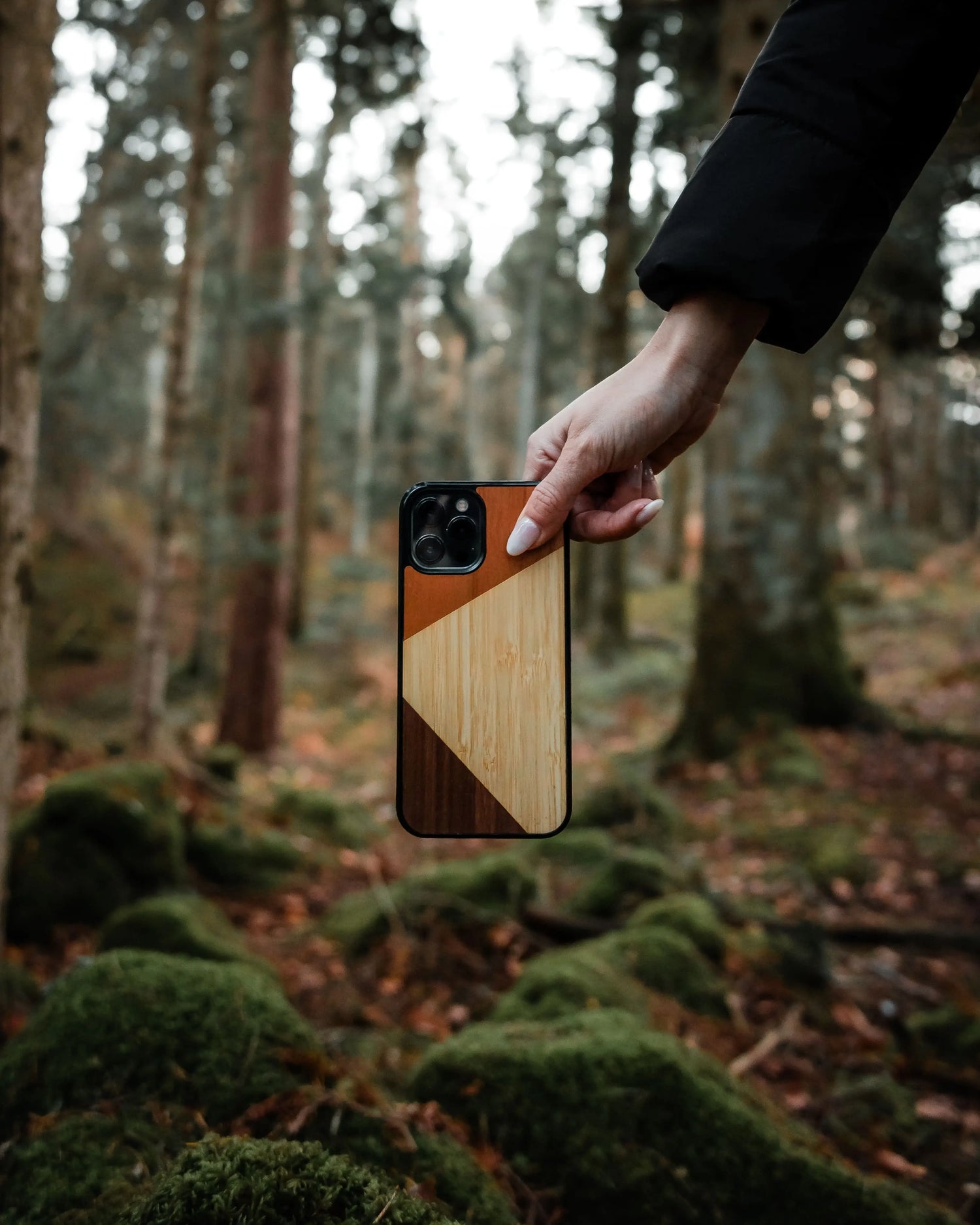 Main dans la forêt qui tient un téléphone avec une coque en bois, artisanale et fabriquée en France, modèle Le Trio, composée de 3 essences de bois, pour iPhone 13 Pro.