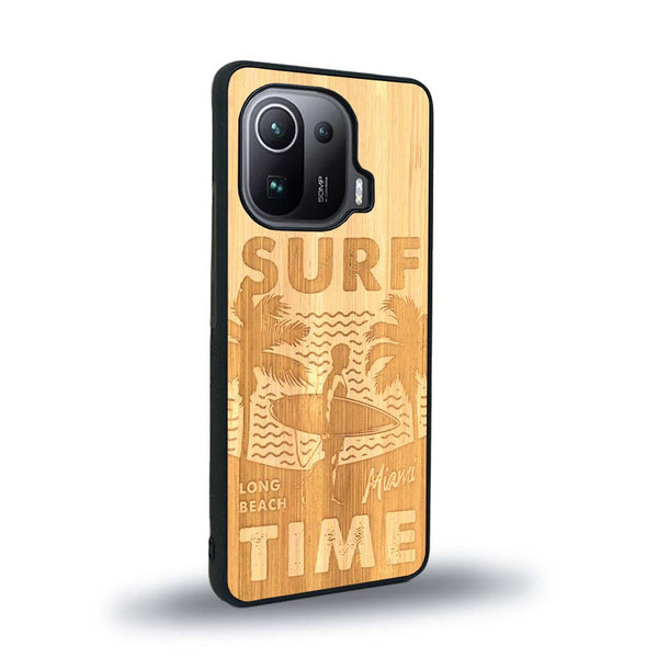 Coque de protection en bois véritable fabriquée en France pour Xiaomi Mi 11 Pro sur le thème chill avec un motif représentant une silouhette tenant une planche de surf sur une plage entouré de palmiers et les mots "Surf Time Long Beach Miami"