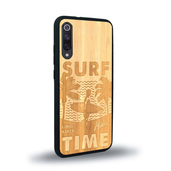 Coque de protection en bois véritable fabriquée en France pour Xiaomi Mi 9SE sur le thème chill avec un motif représentant une silouhette tenant une planche de surf sur une plage entouré de palmiers et les mots "Surf Time Long Beach Miami"